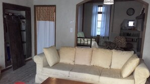 Una sala de estar con un sofá color beige, una mesa de centro de madera y un sillón rojo. Se ve un espejo que refleja una cama y una ventana con cortinas.