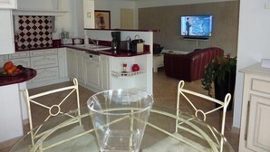 A modern kitchen with white cabinets, a red countertop, and a glass table. There is a TV mounted on the wall and a red leather couch in the living area. - Charming villa with heated pool in quiet, 6/8 people, Gulf of St Tropez (Le Plan-De-La-Tour)