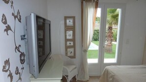 A bedroom with a bed, a television on a dresser, a chair, a door leading to a garden, and wall decorations. - Charming villa with heated pool in quiet, 6/8 people, Gulf of St Tropez (Le Plan-De-La-Tour)