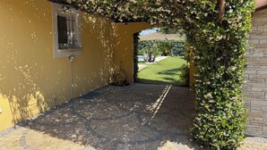 A covered patio area with a yellow wall, a window, and a view of a green lawn. - Charming villa with heated pool in quiet, 6/8 people, Gulf of St Tropez (Le Plan-De-La-Tour)