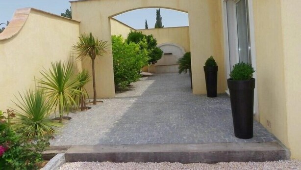 A paved pathway leading to a building entrance with a covered archway, surrounded by potted plants and greenery. - Charming villa with heated pool in quiet, 6/8 people, Gulf of St Tropez (Le Plan-De-La-Tour)