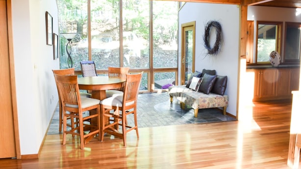 A room with a wooden dining table and chairs, a sofa, and a large window with a view of greenery.