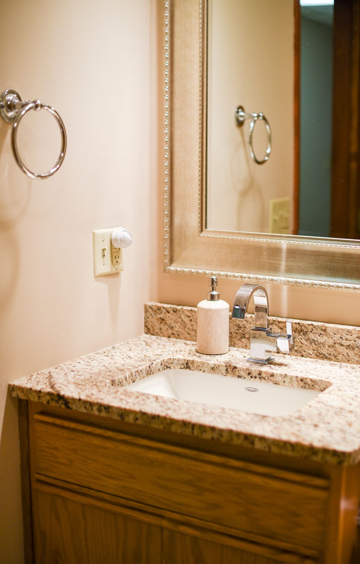 A bathroom with a granite countertop sink, a mirror with a decorative frame, and a towel ring on the wall.