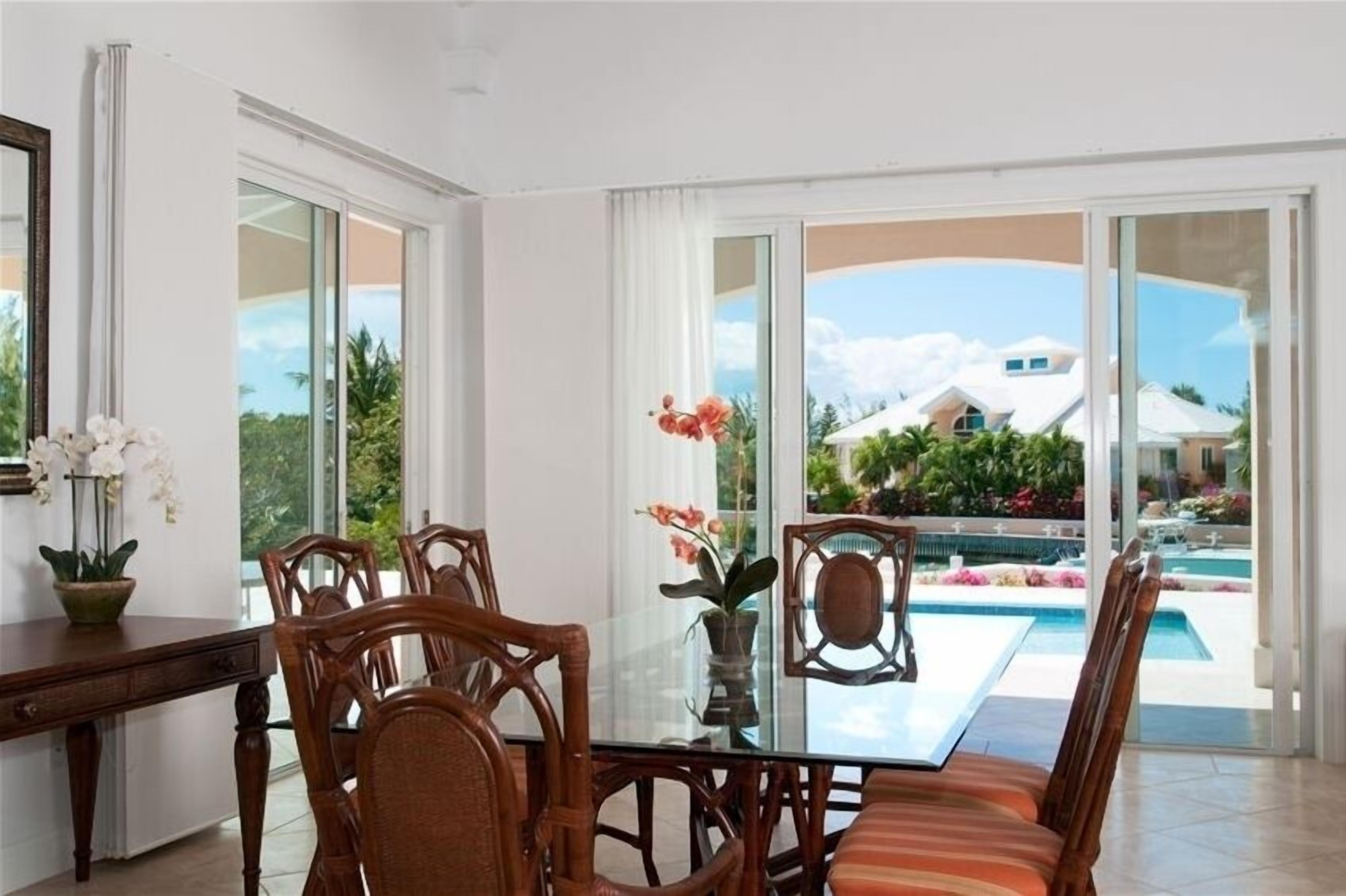 Ein Essbereich mit einem Glastisch und Holzstühlen, einer Vase mit Blumen sowie Blick auf einen Swimmingpool und Gebäude.