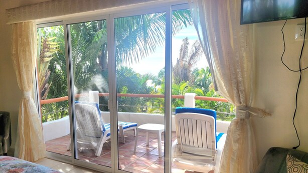 A room with a bed, a TV, a balcony with a chair and table, and a view of palm trees. - Secluded 2 bedroom duplex with private pool and gorgeous jungle and ocean views! (El Divisadero)