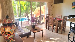 A living room with a sofa, a coffee table, a TV mounted on the wall, a dining area with chairs and a table, and a view of a swimming pool through large windows. - Secluded 2 bedroom duplex with private pool and gorgeous jungle and ocean views! (El Divisadero)