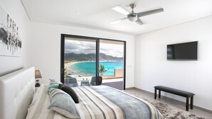 A bedroom with a large bed, a ceiling fan, a TV mounted on the wall, and a view of a beach through a sliding door. - OCEAN FRONT 3 BEDROOM PENTHOUSE UNIT. STAY IN LUXURY. (Beacon Hill,)