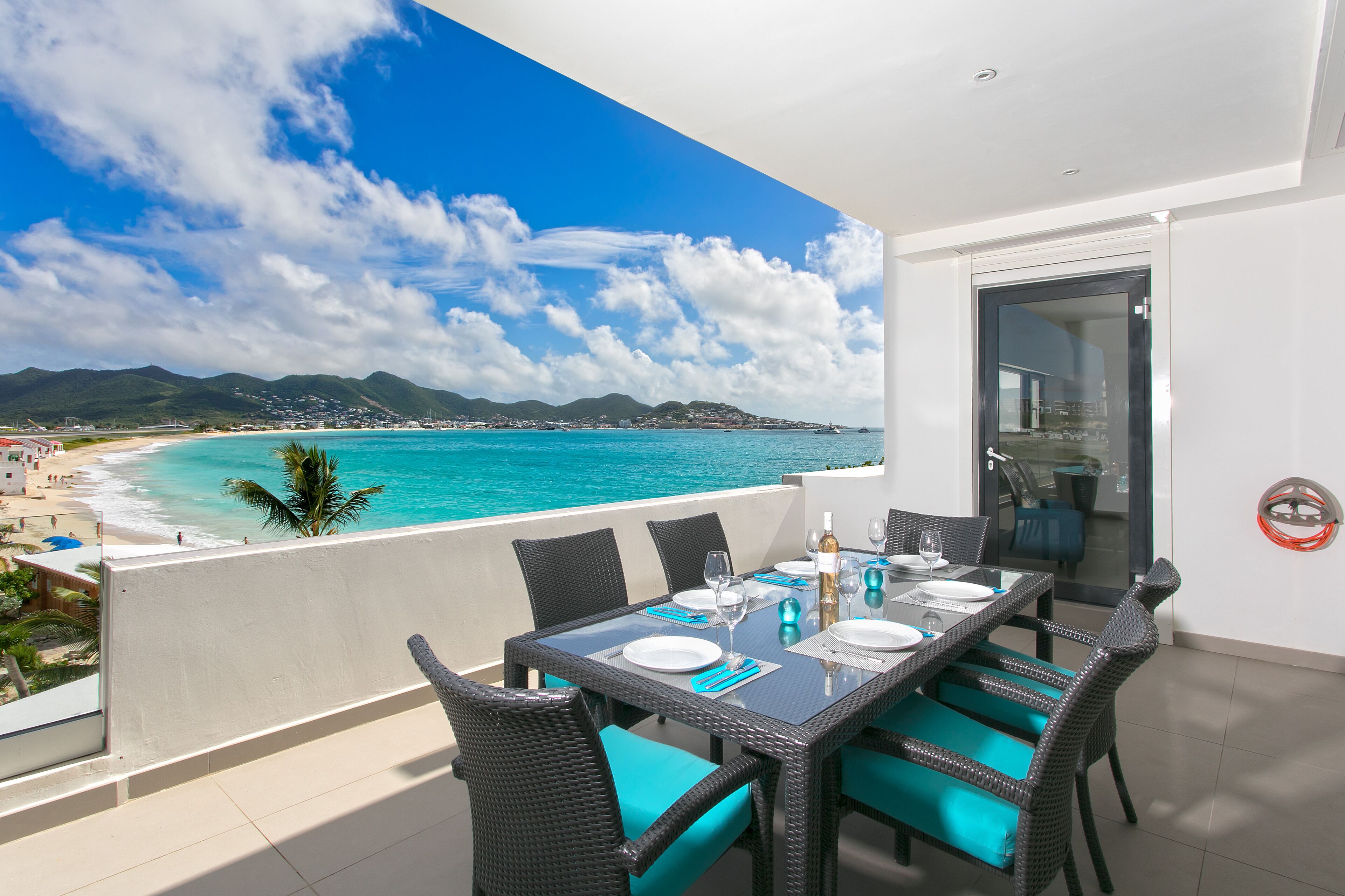 A balcony with a table set for dining, overlooking a beach and turquoise sea.