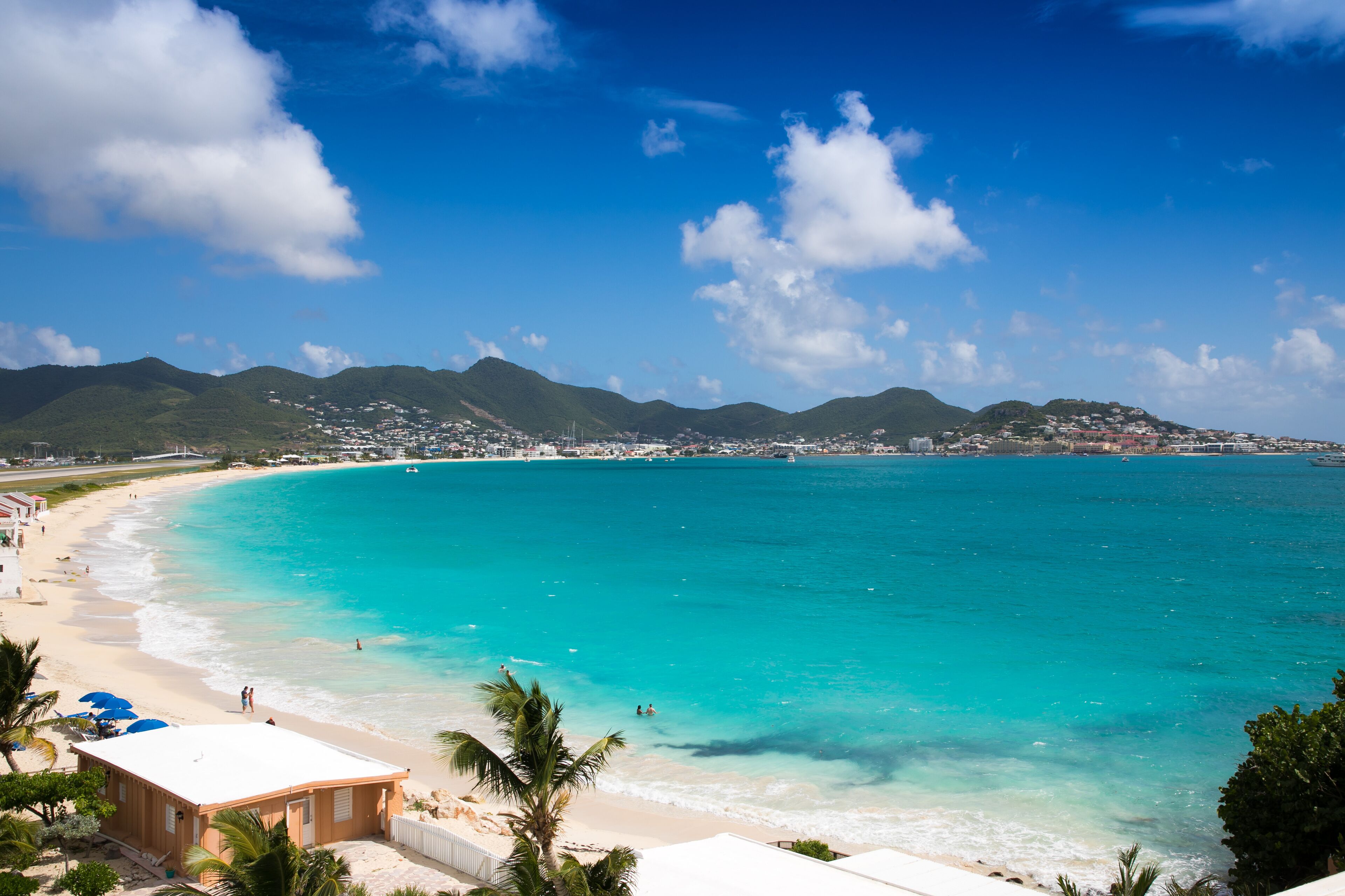 Vista costeira com praia, águas turquesa cristalinas e uma cidade ao fundo.