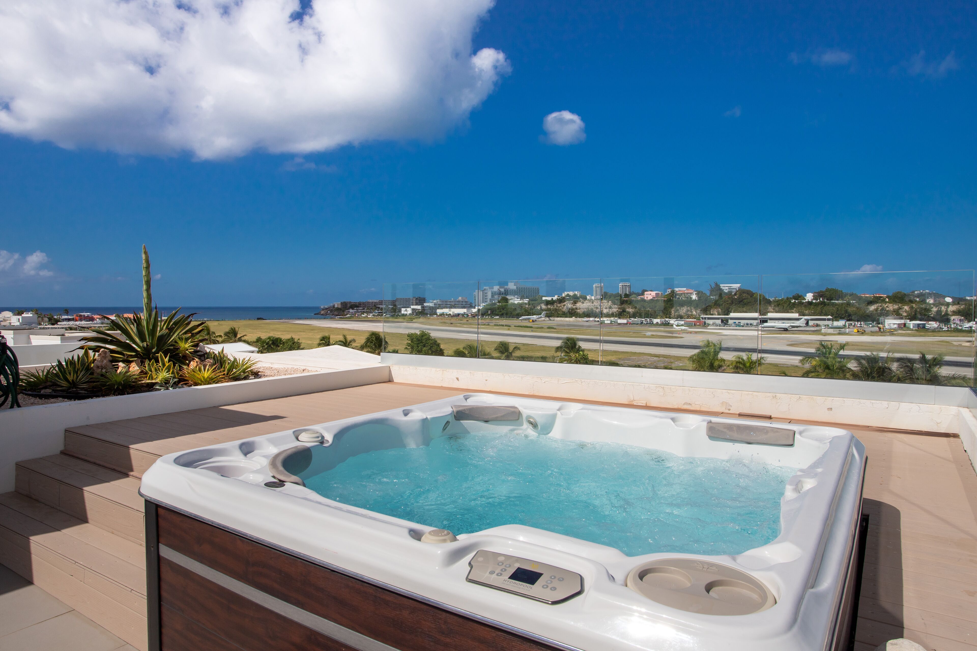 A hot tub with a control panel, surrounded by a wooden deck and plants, overlooking a cityscape and the ocean.