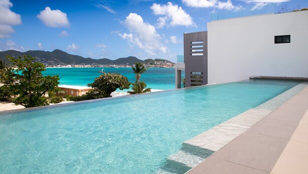 A modern pool with clear blue water, surrounded by a white building and a view of the sea and mountains. - OCEAN FRONT 3 BEDROOM PENTHOUSE UNIT. STAY IN LUXURY. (Beacon Hill,)