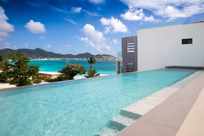 Una piscina moderna con aguas azules cristalinas, rodeada por un edificio blanco y vistas al mar y las montañas.
