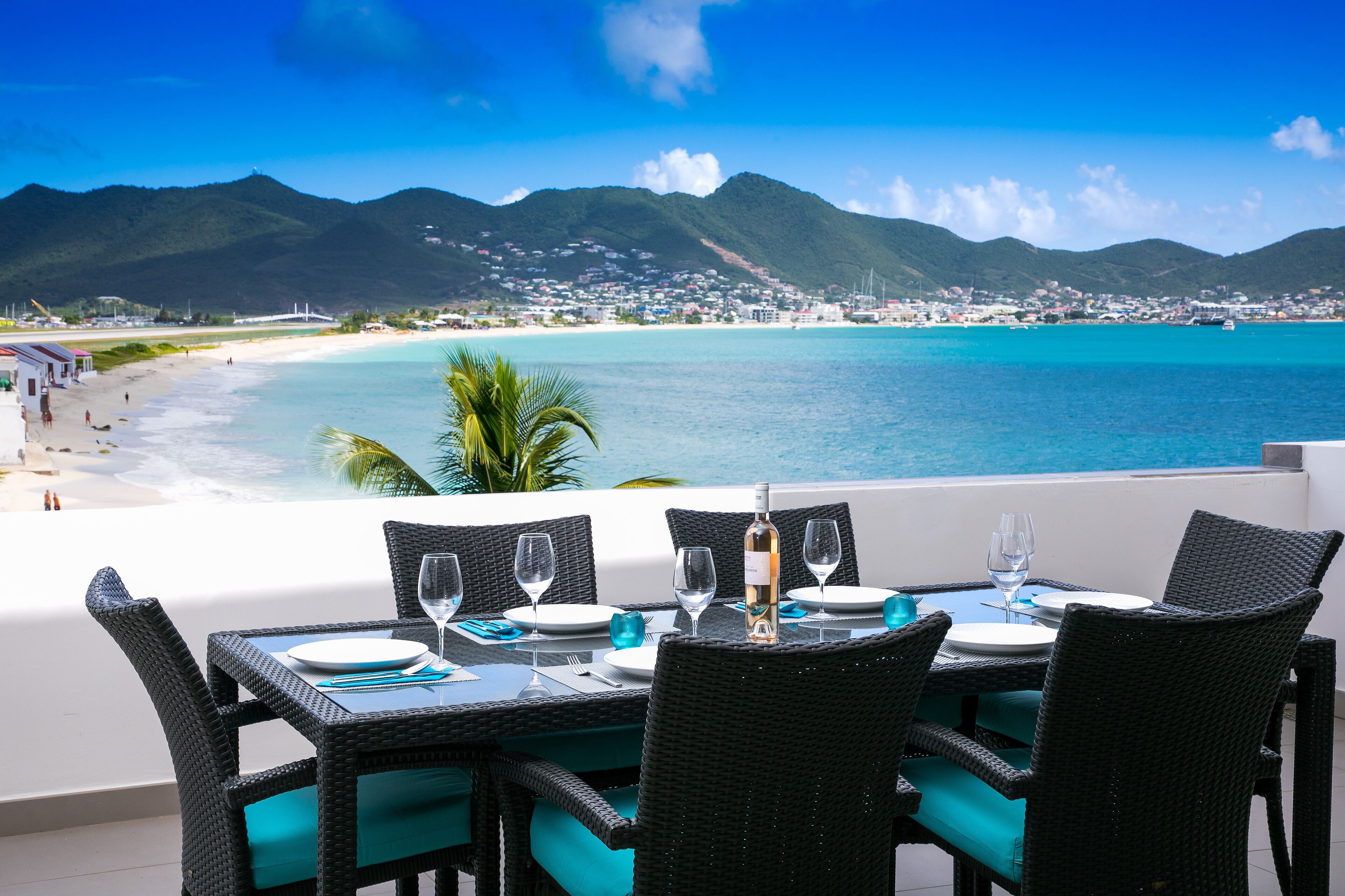 A dining table set with chairs, a bottle, and glasses, overlooking a beach and turquoise waters with mountains in the background.