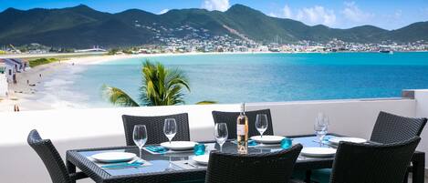 A dining table set with chairs, a bottle, and glasses, overlooking a beach and turquoise waters with mountains in the background.