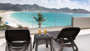 A balcony with two chairs and a table, overlooking a beach with turquoise water and mountains in the background. - OCEAN FRONT 3 BEDROOM PENTHOUSE UNIT. STAY IN LUXURY. (Beacon Hill,)