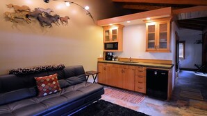 A cozy living area with a sofa, a small table, and a kitchen with wooden cabinets and a microwave. - Carriage House  - Tecumseh's timber frame Cottage (Anchorage)