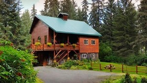 A rustic wooden cabin with a green roof, a porch with railings, and a staircase leading to the upper level. - Carriage House  - Tecumseh's timber frame Cottage (Anchorage)
