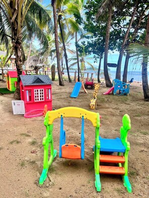 A beach with a swing set, a small red house, and a giraffe-shaped slide.