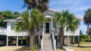 A two-story house with a porch, a flag, and palm trees. - The Gulls Nest Beach House (Edisto Island)
