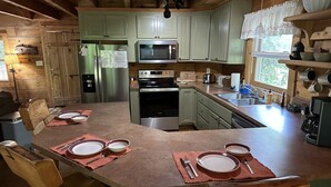 A wooden cabin-style kitchen with a central island, stainless steel appliances, and a window with a view of trees. - Peaceful Real Log Cabin Rushing Trout Stream  Jacuzzi Hot Tub (deep gap)