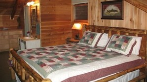 A wooden cabin bedroom with a bed, a nightstand, a lamp, a mirror, and a framed picture on the wall. - Peaceful Real Log Cabin Rushing Trout Stream  Jacuzzi Hot Tub (deep gap)