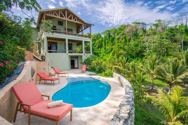 Une maison à deux étages avec une piscine, entourée d’une végétation luxuriante et de palmiers.