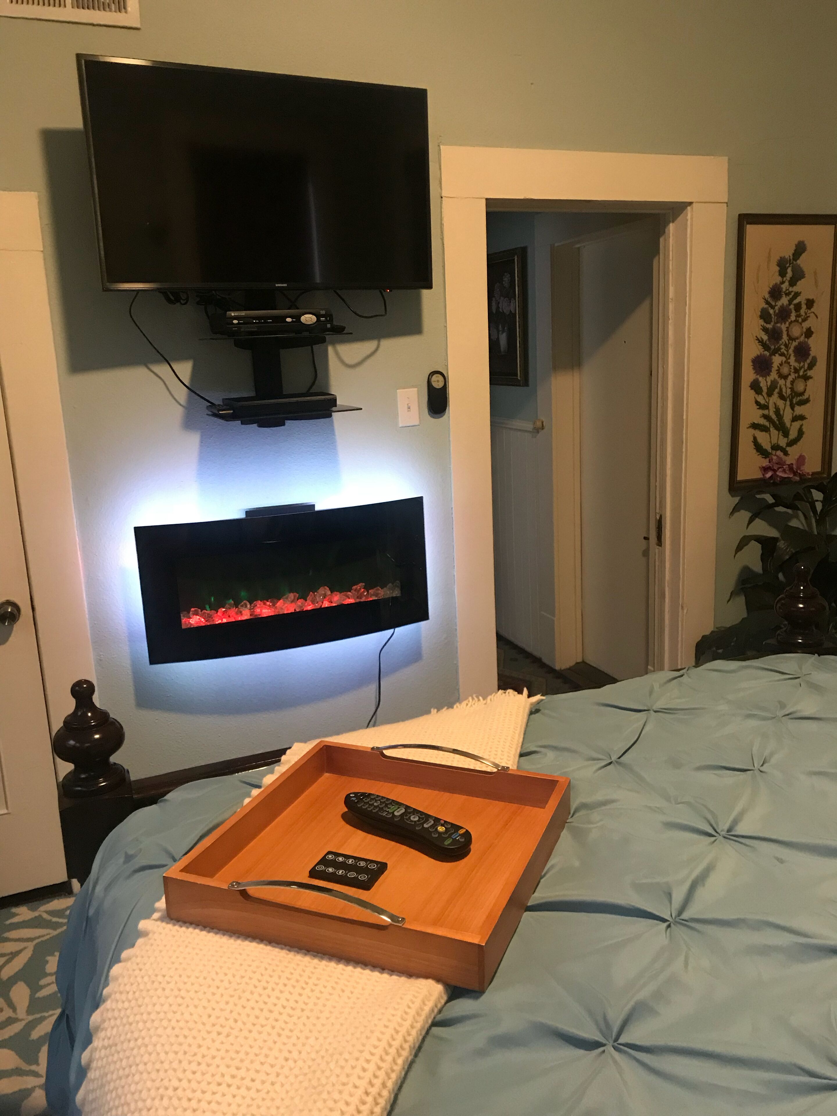 Un dormitorio con televisor montado en la pared, una mesita de noche con una bandeja que sostiene un control remoto, y una cama con colcha azul.