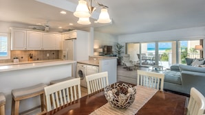 A modern kitchen with white cabinets, a dining area with a wooden table and white chairs, and a living room with a gray sofa and a view of the ocean. - Prime Beachfront with Views Views Views! (Siesta Key)