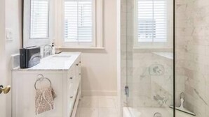 Een badkamer met een wit wastafelmeubel, een douche met een glazen deur en een raam met zonwering.