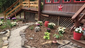 Une terrasse en bois avec un escalier menant à un espace abrité, le tout entouré d’un jardin où poussent différentes plantes et fleurs.