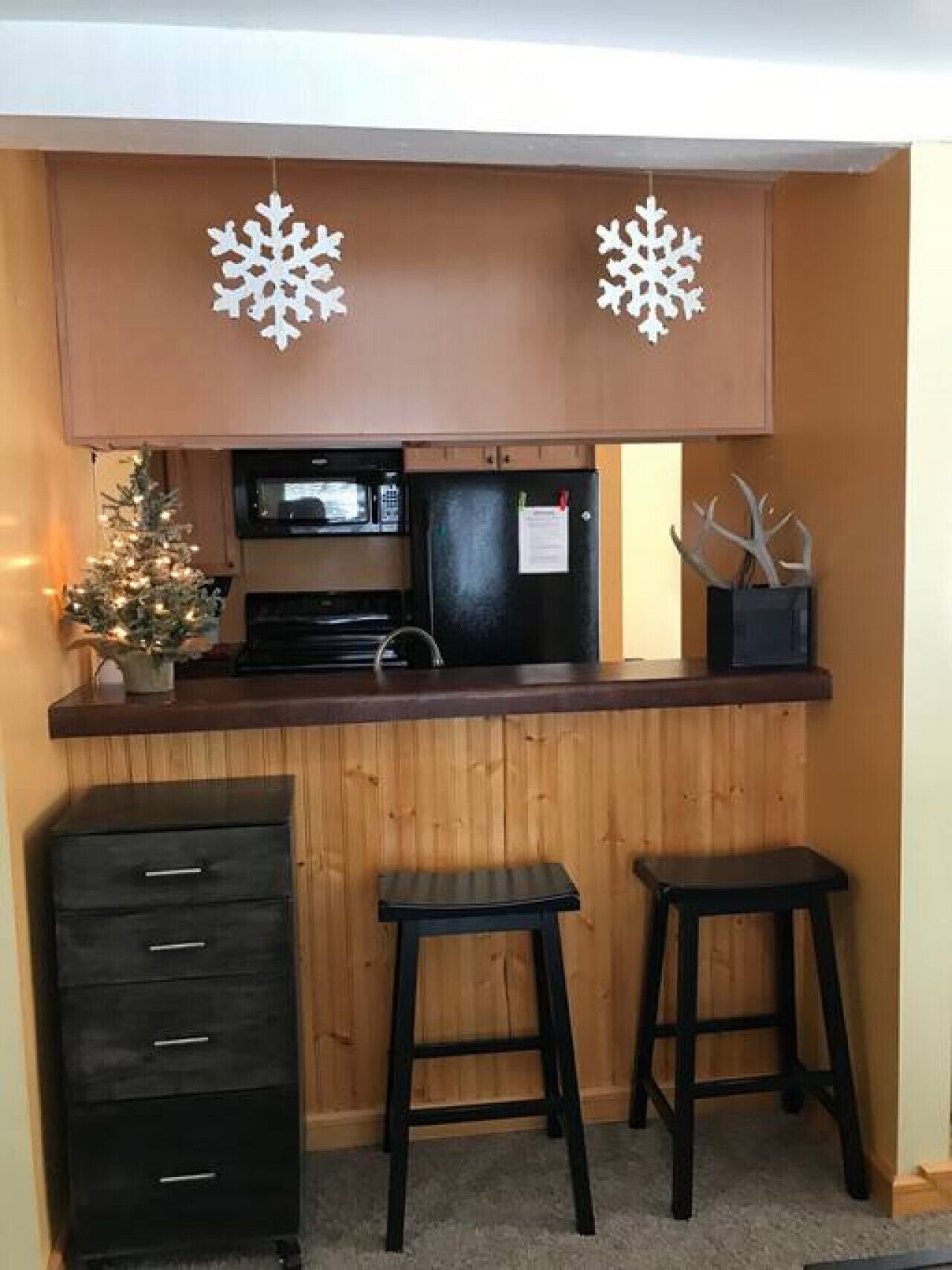 Une cuisine compacte avec des meubles en bois, un réfrigérateur noir, un four à micro-ondes et un petit sapin de Noël. On y trouve deux tabourets et un meuble noir avec des tiroirs.