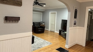 Una sala de estar con piso de madera, un tapete gris, un sillón de cuero negro, un ventilador de techo y un televisor.