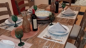 Una mesa de comedor dispuesta con platos, vasos y una botella de vino, rodeada de sillas y un sofá al fondo.