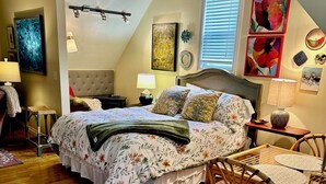 A bedroom with a bed, a nightstand, a lamp, a chair, a table, and a window with blinds.