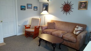 A living room with a beige sofa, a wooden armchair, a coffee table, and a floor lamp. There are framed pictures on the wall and a sun decoration. - Quiet, clean, and comfy: Centrally Located In The Mt. Washington Valley. (Intervale)