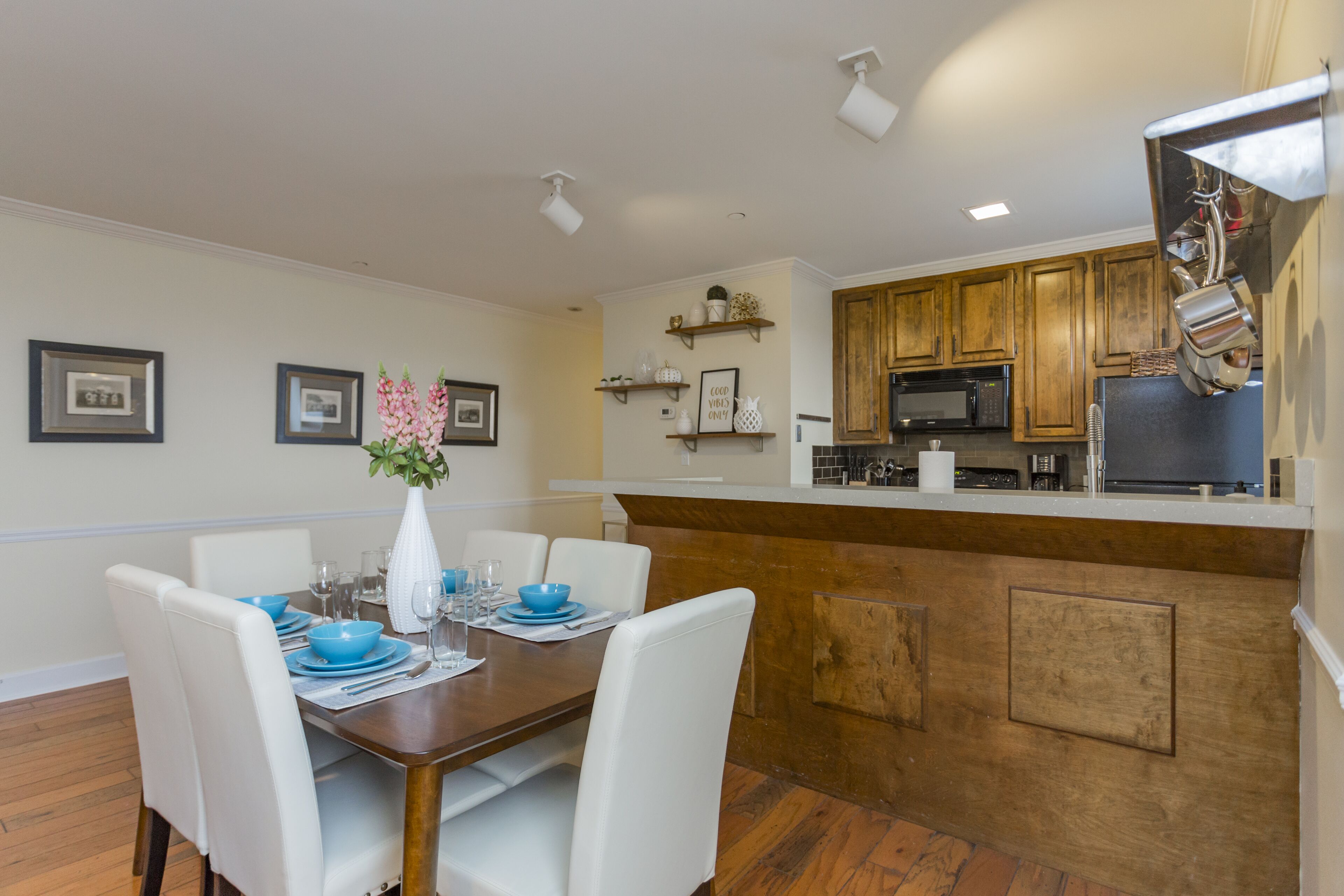 Un comedor con una mesa de madera, sillas blancas y cuencos azules. Se ve una cocina con armarios de madera y un microondas.