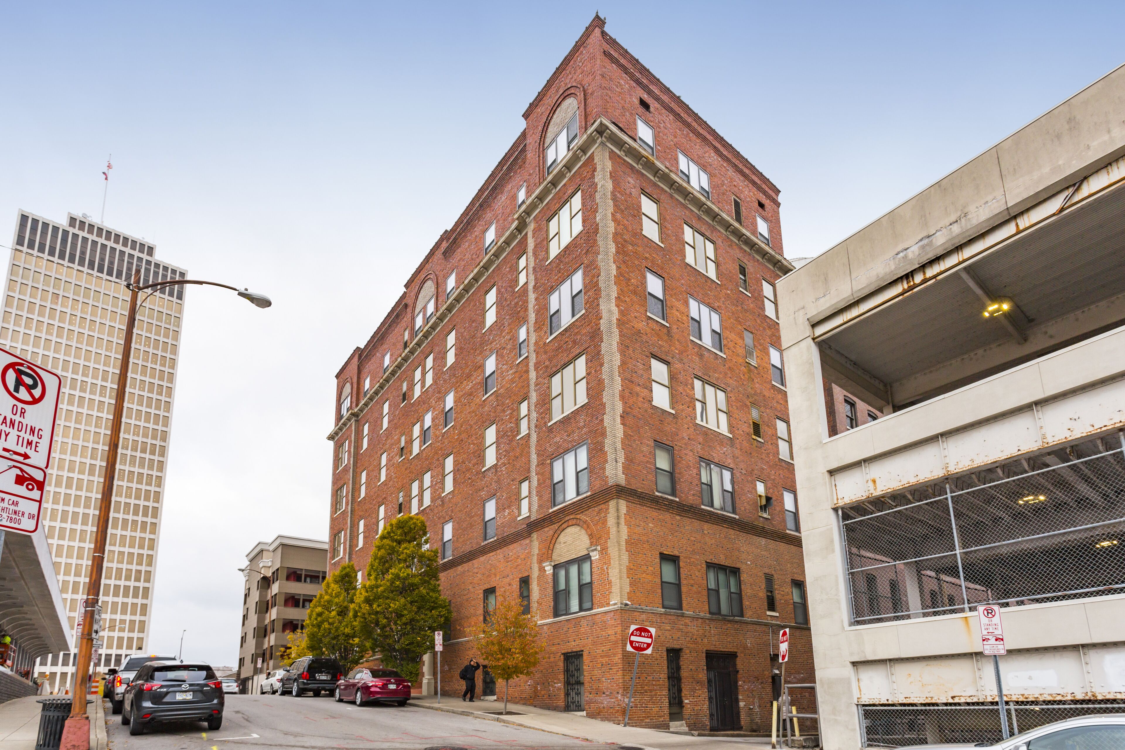 A multi-story brick building with a corner entrance, a parking sign, and a street view with cars and a pedestrian.