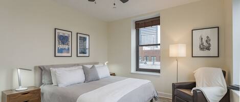 A bedroom with a bed, a nightstand, a chair, a ceiling fan, and a window with blinds.