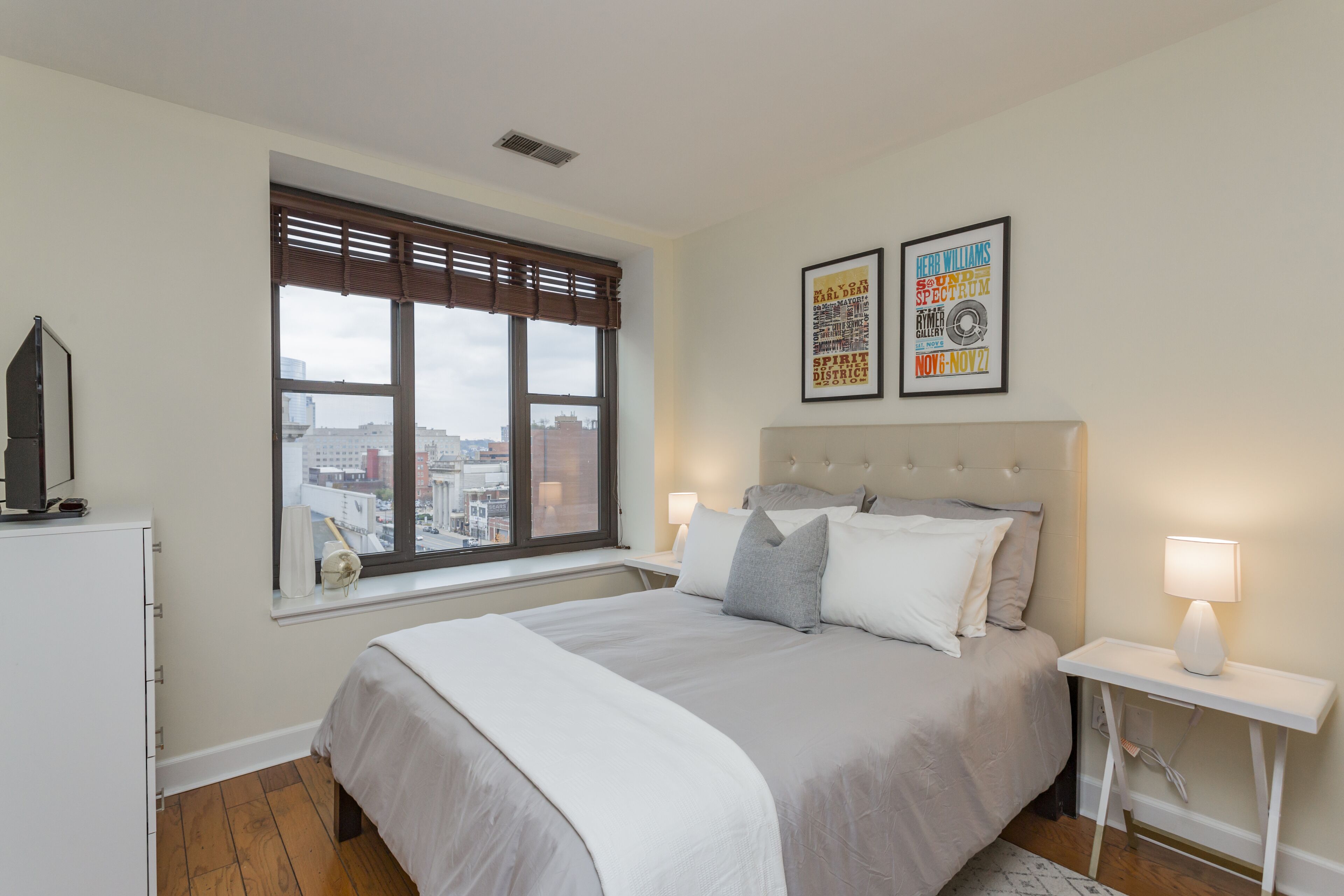 A bedroom with a large bed, a window with blinds, a bedside table with a lamp, and two framed pictures on the wall.