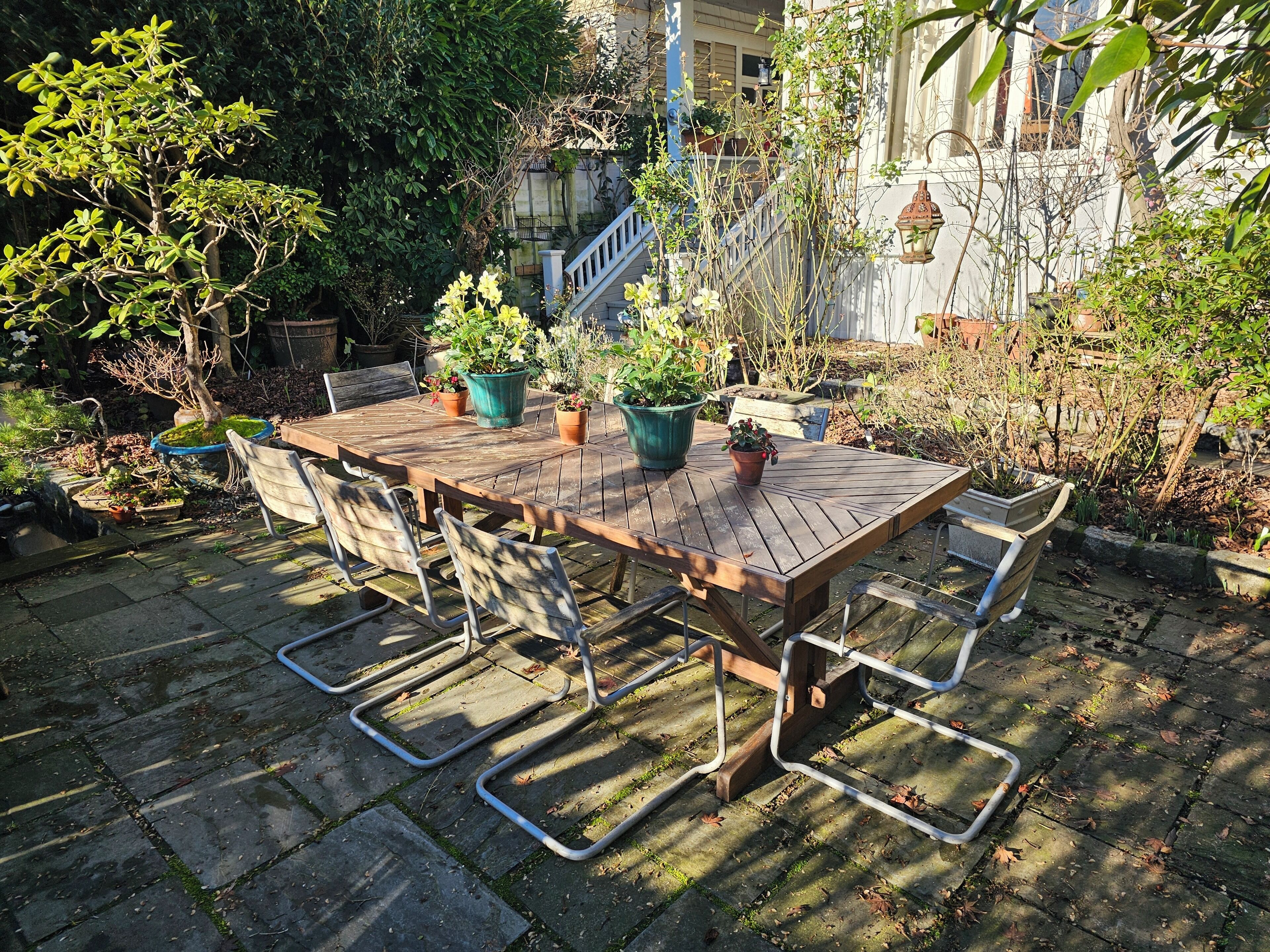An outdoor wooden table with metal chairs, surrounded by potted plants and a stone-paved area.