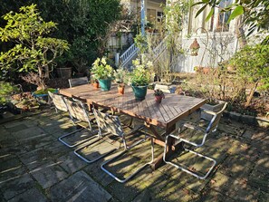 An outdoor wooden table with metal chairs, surrounded by potted plants and a stone-paved area. -  Gracious and spacious craftsman house seaside and cityside in Kitsilano. (Vancouver)