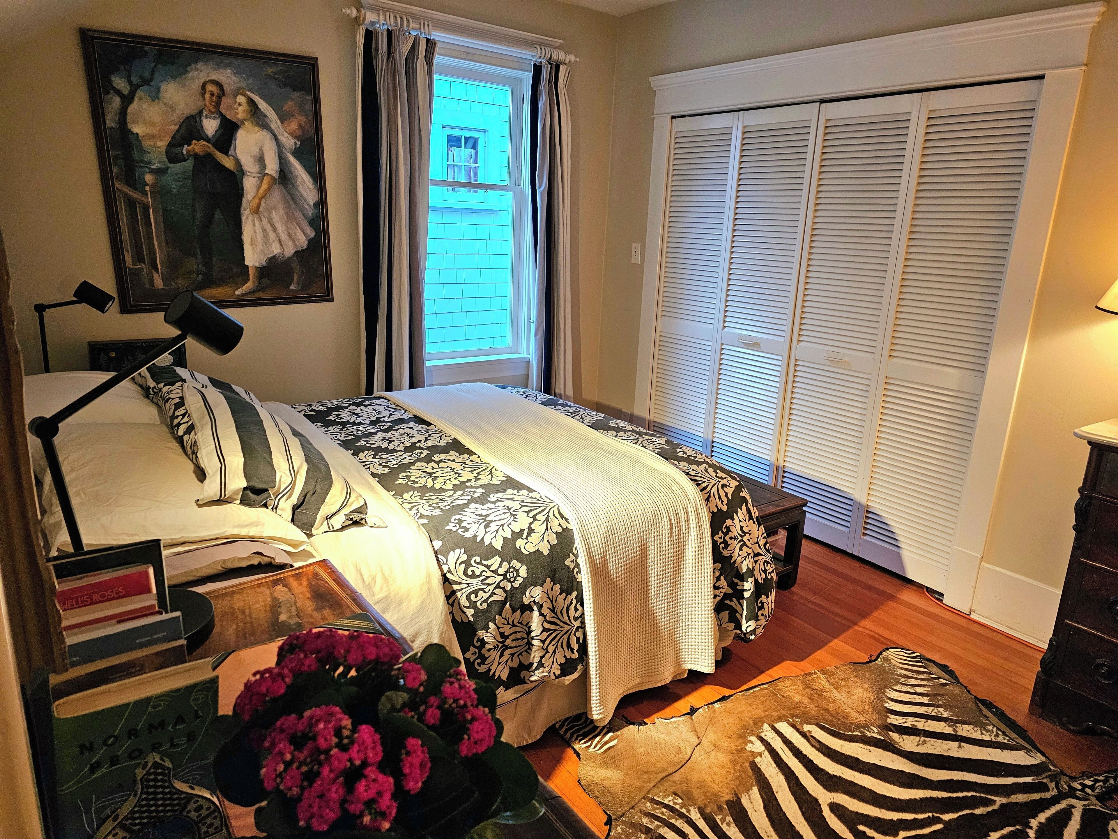 Un dormitorio con una cama, una mesita de noche con libros, una lámpara, una ventana con cortinas, un cuadro y una puerta corredera para el armario.