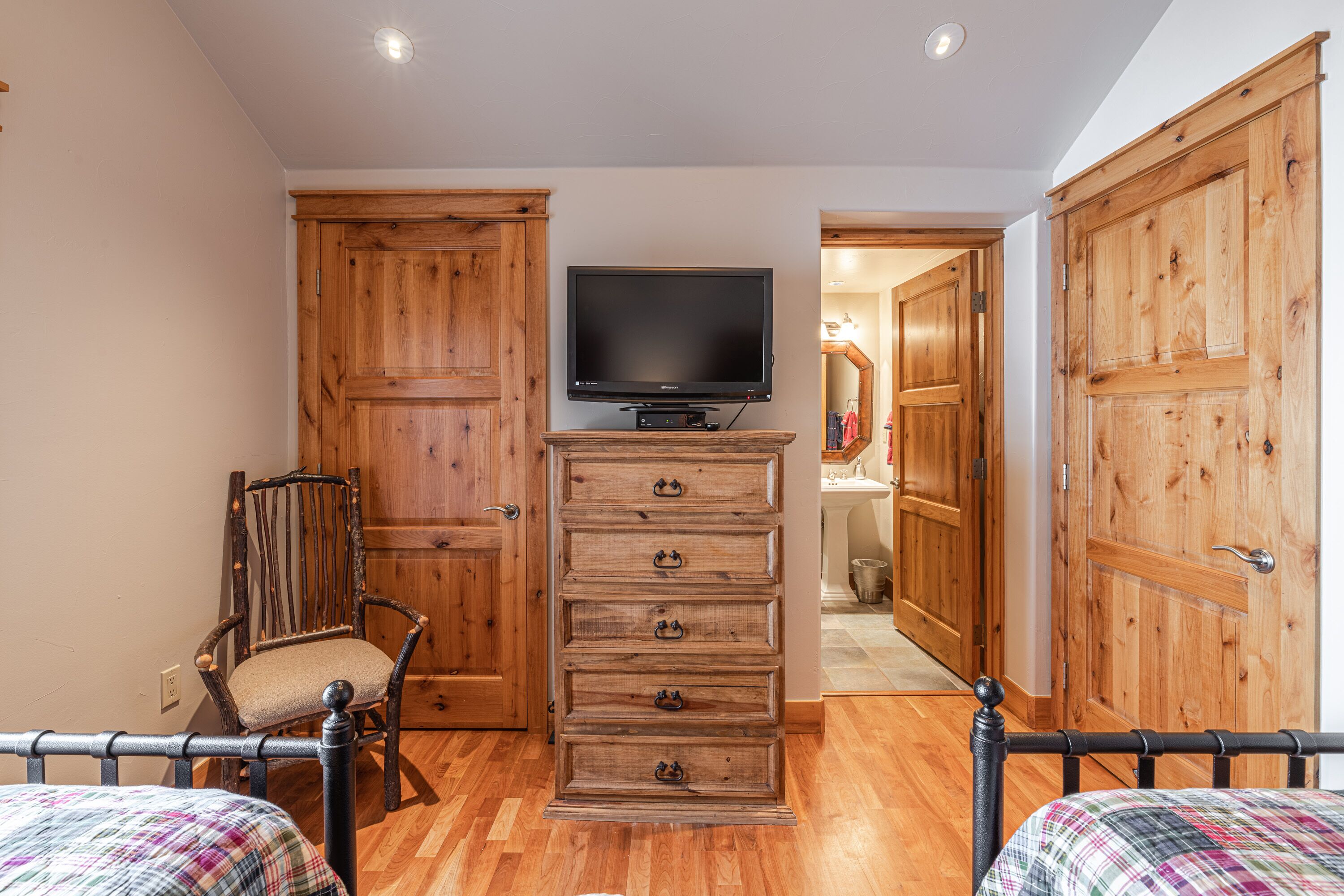 A room with a wooden dresser, a television on top, a chair, and a bed with a plaid bedspread.
