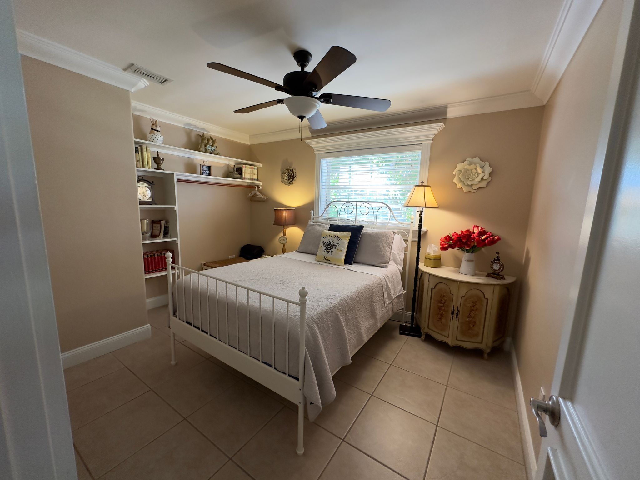 A bedroom with a white bed, a ceiling fan, a window with blinds, a bedside table with a lamp, and a shelf with books and decorative items.