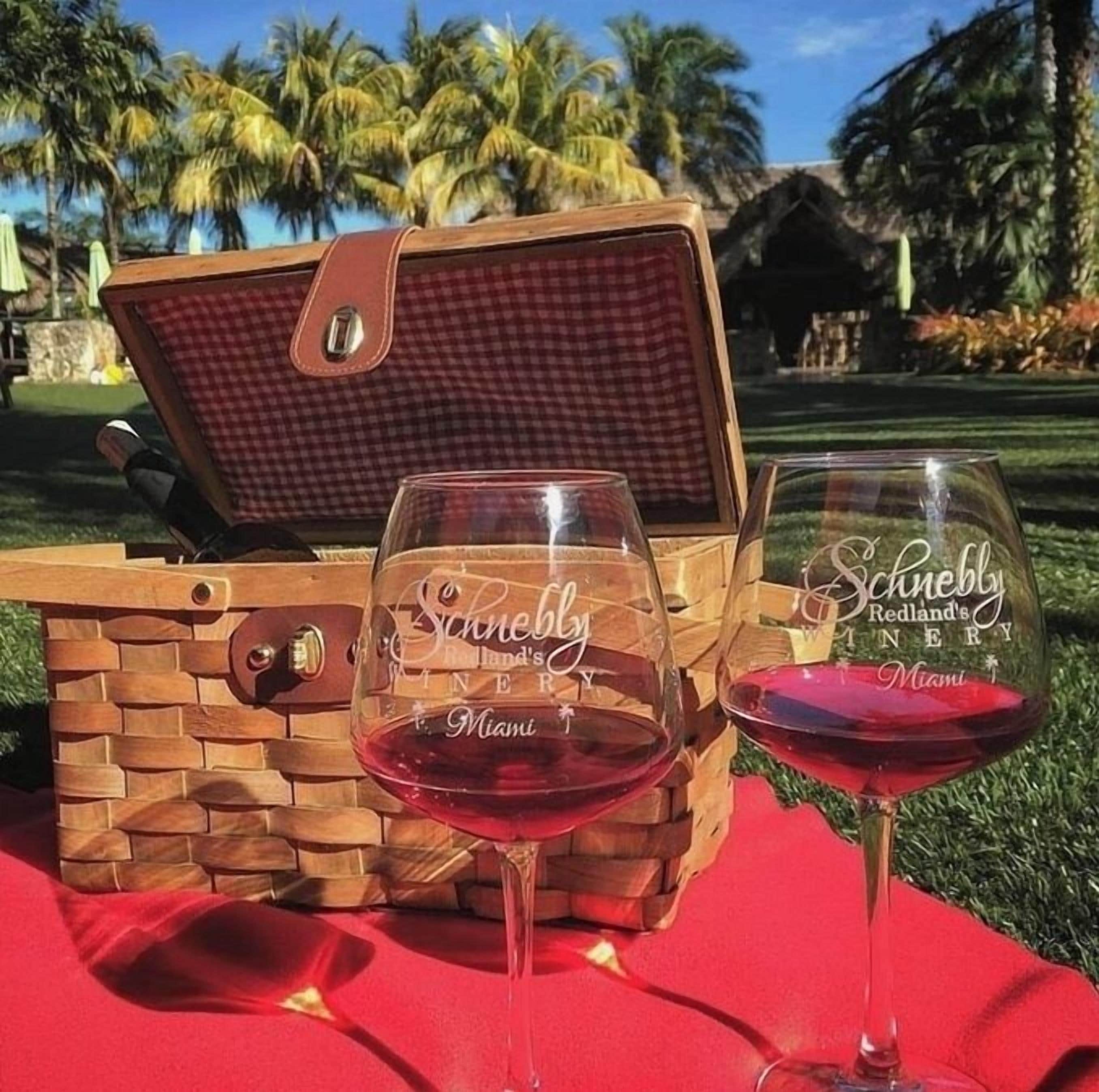 A picnic setup with a wooden picnic table, a checkered picnic basket, and two wine glasses with 'Schnebly Redland's WINERY Miami' etched on them.
