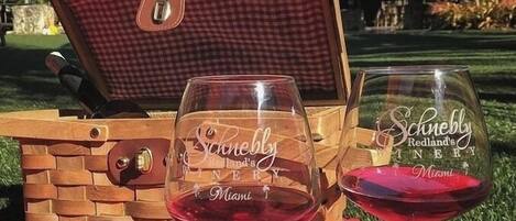 A picnic setup with a wooden picnic table, a checkered picnic basket, and two wine glasses with 'Schnebly Redland's WINERY Miami' etched on them.