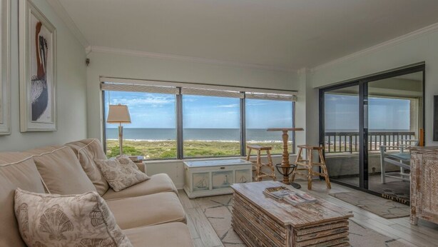 A spacious living room with a large window overlooking the ocean, a beige sofa, a wooden coffee table, and a view of the beach through the glass door. - Beautiful Oceanfront Condo With Breathtaking Beach Views , 3RD floor, C117 (Fernandina Beach)