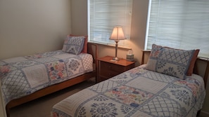 Une chambre à coucher avec deux lits simples, chacun recouvert d’une courtepointe à motifs, une table de chevet avec une lampe et une fenêtre équipée de stores.