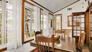 Comedor con mesa de madera, sillas y un tazón. Hay ventanas con cortinas, techo de madera y vistas al exterior con vegetación.