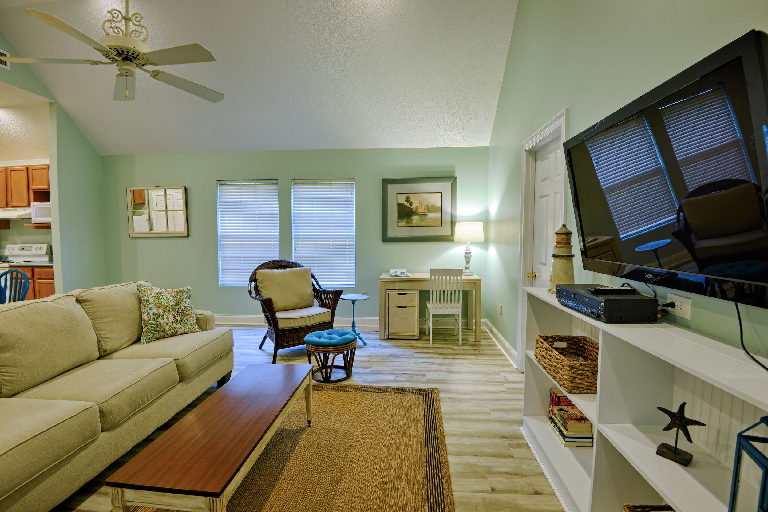 A living room with a sofa, coffee table, and a TV on a stand. There is a desk with a chair, a window with blinds, and a bookshelf.